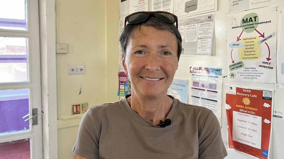 Rona wearing a grey T-shirt sits indoors in front of a noticeboard covered with community and recovery support posters. A small microphone is attached to the shirt, and sunglasses rest on the person's head. A glass-panelled door is visible to the left.