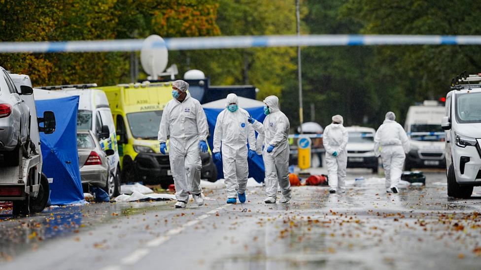 People walks in hazmat suits - their faces covered with surgical masks and hands covered in blue gloves - walk down a road blocked off by police tape and lined with emergency vehicles