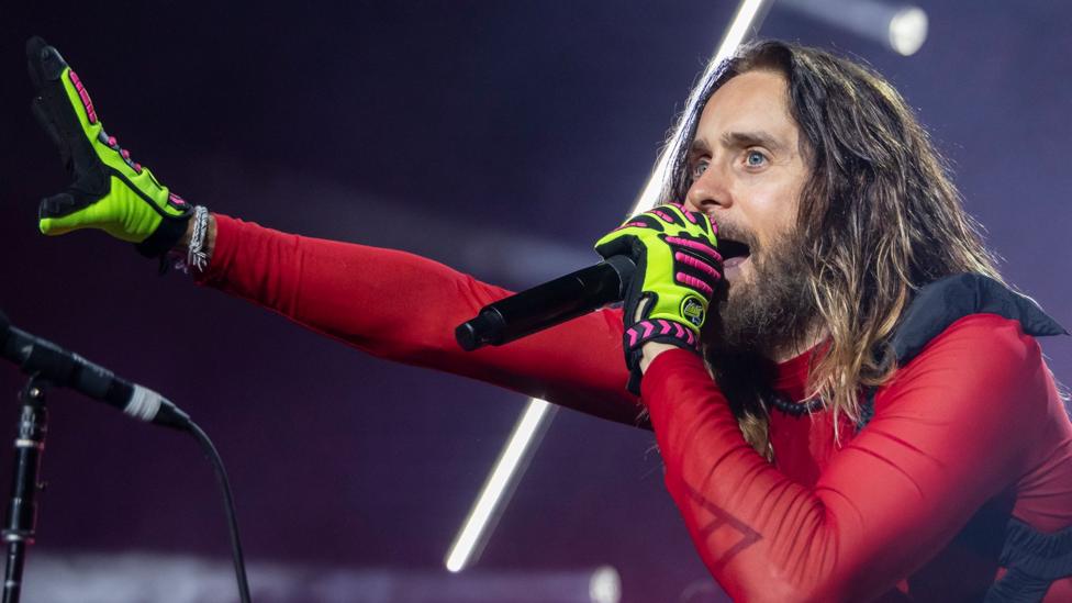 Jared Leto stands under concert lighting, singing into a handheld microphone. He has long hair and a beard and is dressed in a red, sheer and fitted top with neon green and pink gloves. One arm is stretched outward toward the audience while the other holds the microphone close to his mouth. Bright white stage lights form diagonal lines in the background. CLARKSTON, MICHIGAN - AUGUST 06: Actor/Singer Jared Leto of Thirty Seconds To Mars performs during their Seasons World Tour 2024 at Pine Knob Music Theatre on August 06, 2024 in Clarkston, Michigan. (Photo by Scott Legato/)