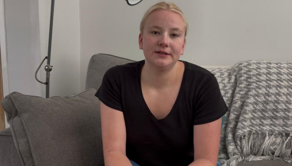 Lowri looks at the camera. She wears a black t-shirt and her blonde hair is tied back. She sits on a grey sofa with a grey and white woollen blanket behind her. 