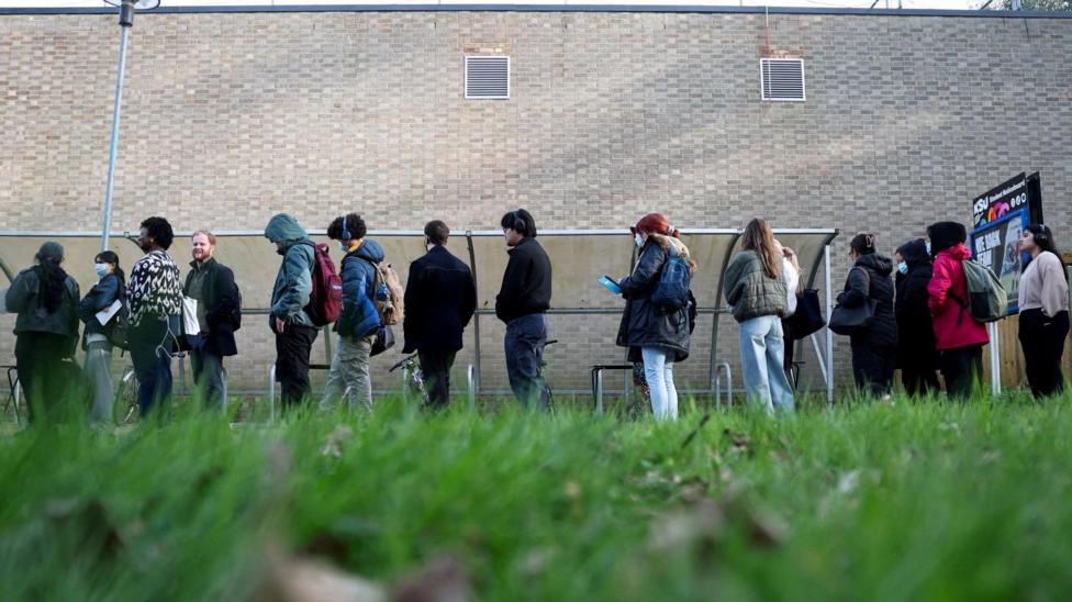 Students queue outside a light-coloured brick building. They are wearing coasts and backpacks, and some of them are wearing face masks. The image is shot from the grass and looking up.