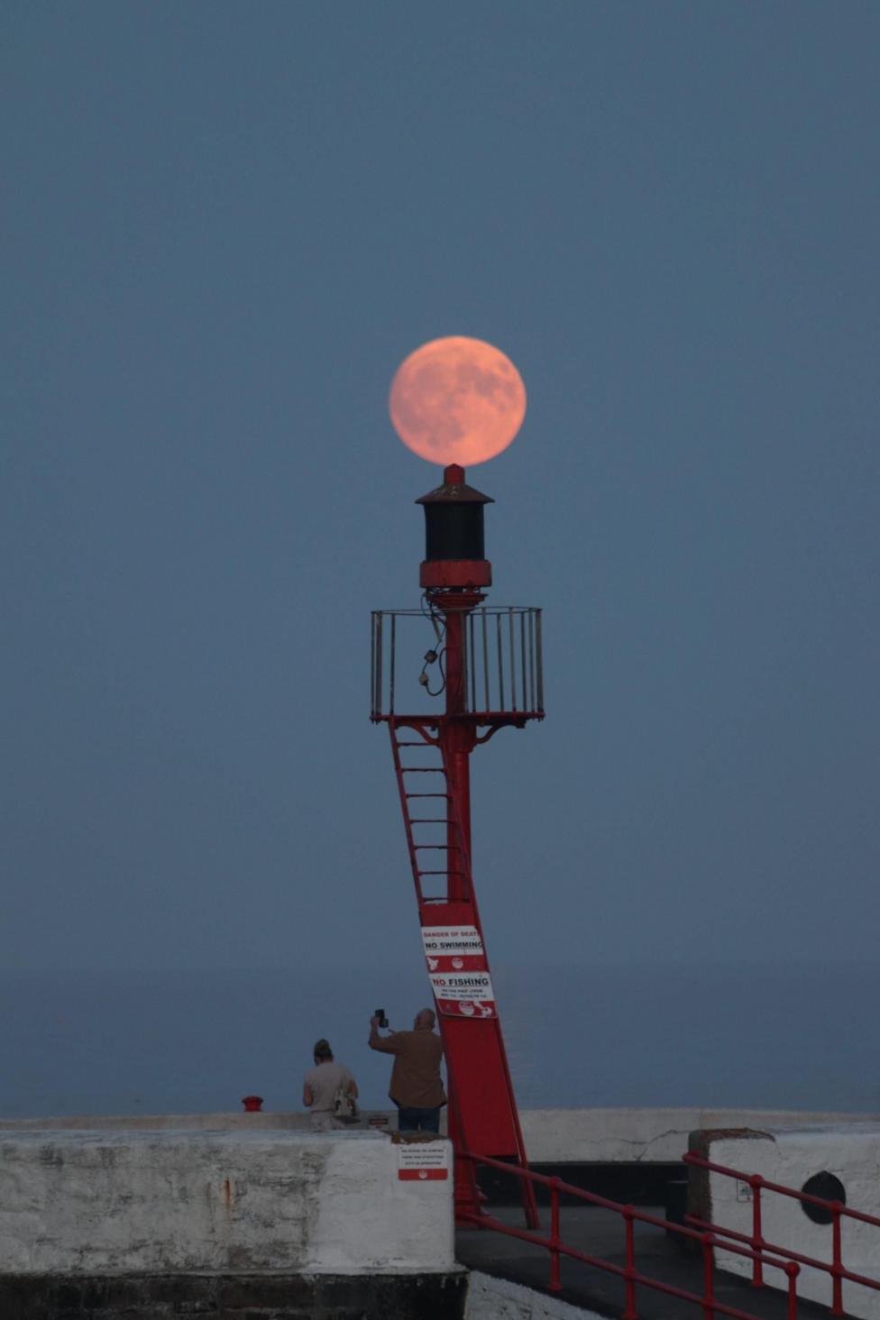 A red harbour navigation beacon stands on a pier in Looe, Cornwall, with a pink-orange full moon appearing directly above it and two people silhouetted nearby.