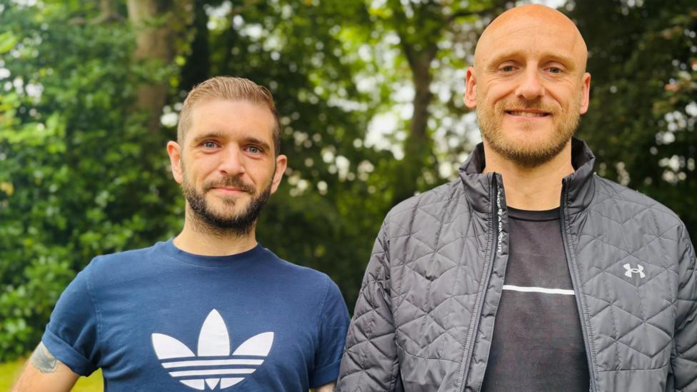 Kieran Hutcheon and Dan Bickmore, two men in their 30s smiling at the camera, in front of trees.