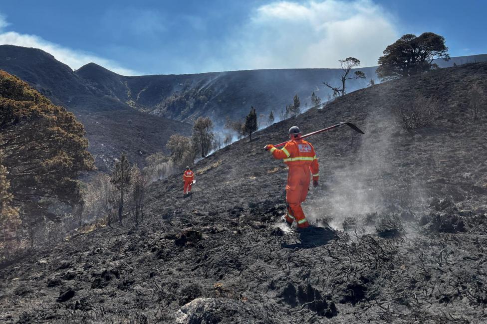 Two people wearing bright orange overalls walk across a scorched, smoking hillside.
