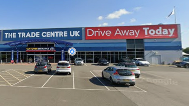 Cars are parked in front of a large windowed store front as part of a larger building. There is a blue sign top left with white writing saying