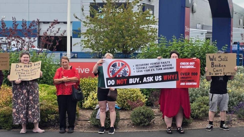 Seven people, a mixture of men and women, stand on the pavement outside a large building belonging to Trade Centre UK. They are holding signs which include the words