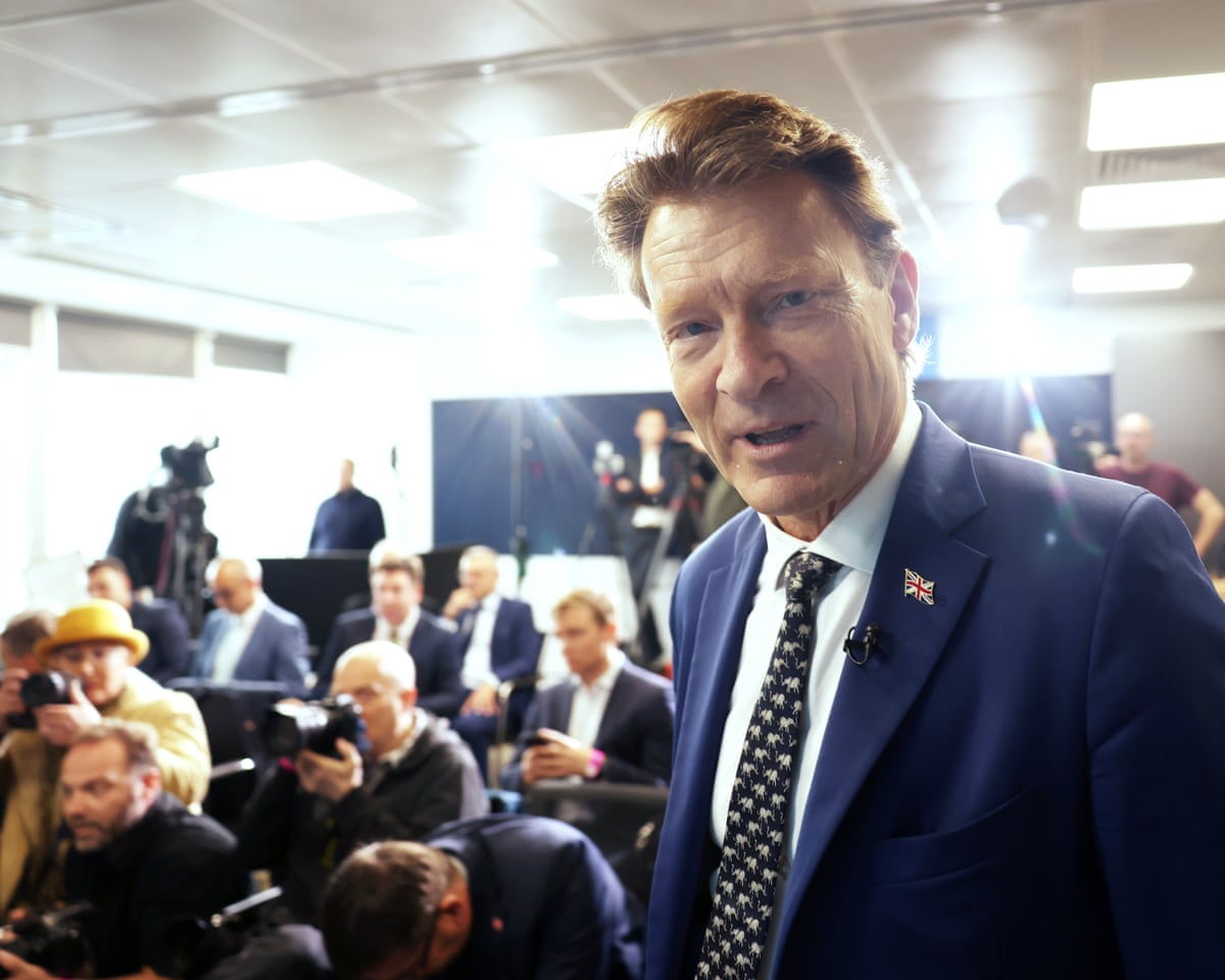 Richard Tice standing up with a number of people and photographers seated behind him.