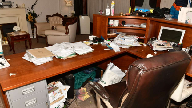 A pristine-looking area with armchairs and flowers contrasts with a messy desk. Under the desk, hidden from view, are various papers and carrier bags.