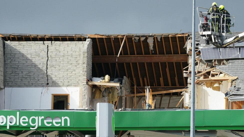 Shows an Applegreen service station with a hole in the rood and members of the emergency services looking down on it to the right from an extending ladder.