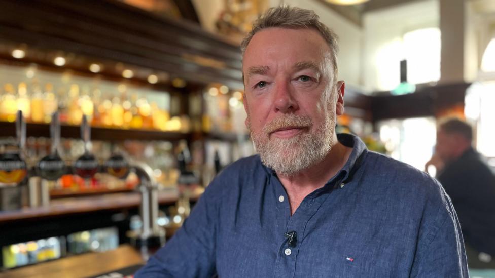 Alistair Brown looking straight at the camera. He has short grey hair and a white beard. He is wearing a blue shirt and is leaning on a bar with taps in the background.