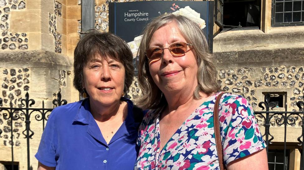 Richard Laversuch's sisters, Cathy Laversuch (short dark hair) and Bridget Ryan (shoulder-length hair and floral dress) pose for a photo in front of the coroner's court, a historical building with ornamental railings.