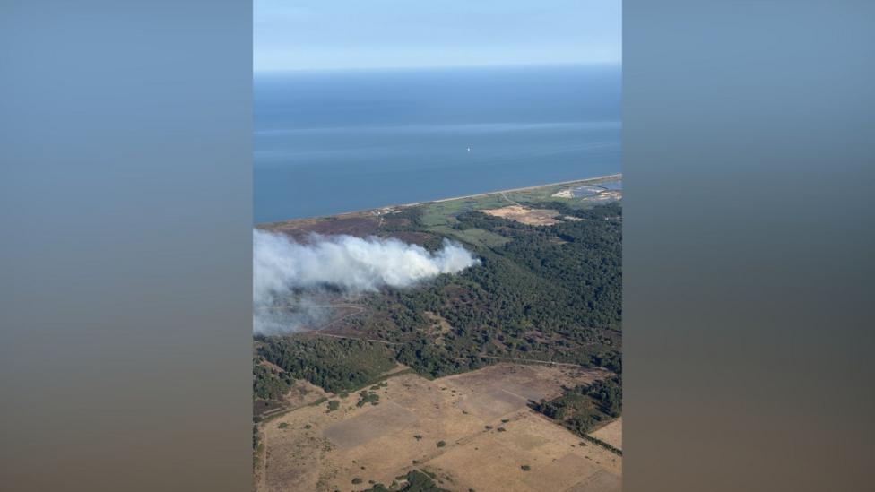 In a picture taken from the air, smoke is drifting from right to left from a wooded area. A large area of open ground is in the foreground, and the sea is at the top of the picture.