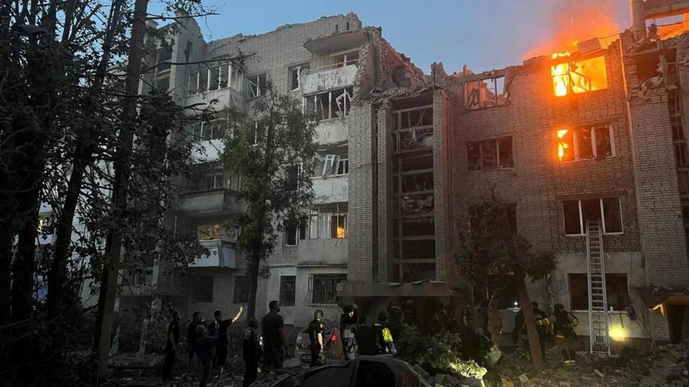 Firefighters work at a site of a building damaged by a Russian missile strike, amid Russia's attack on Lviv, Ukraine. A building is damaged and on fire, with firefighters standing in the foreground. 