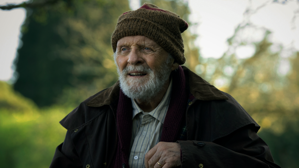 Sir Anthony Hopkins with a grey beard. He wears a dark coat, woollen brown hat and a beige shirt with a blue stripes.