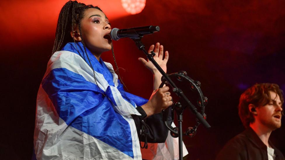 Brooke Combe performing with a Scotland saltire flag over her shoulders. The singer is on stage singing into a microphone.