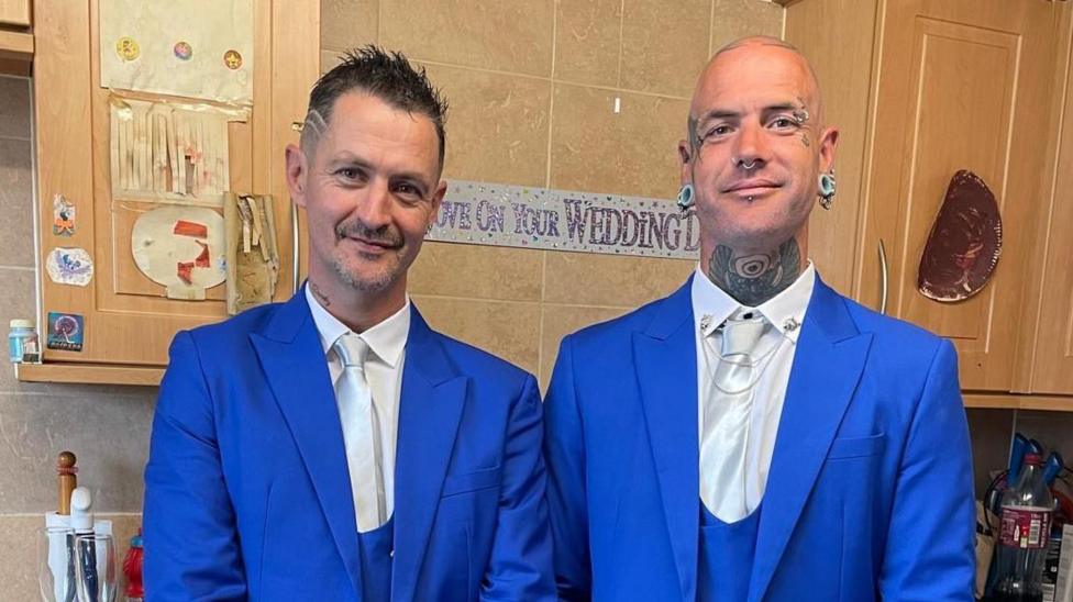 Both men are standing in a kitchen with pale pine cabinets wearing bright blue suits and white shits and tie. Behind them is a sign on the wall that appears to say