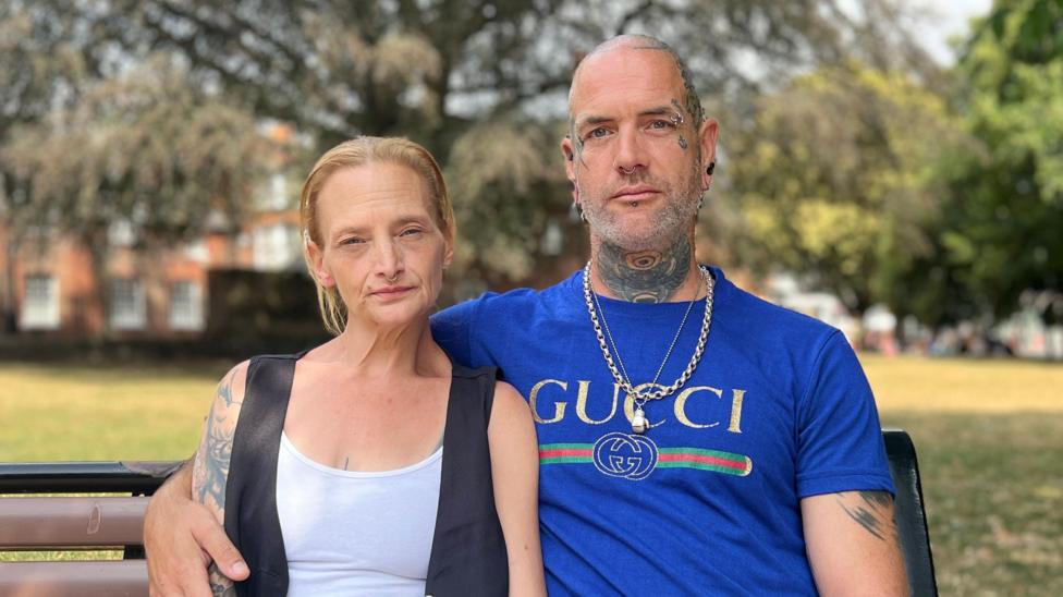 Rebecca and Tony Nye are sitting on a bench with trees and grass visible but not in focus behing. Tony has his arm around Rebecca and is wearing a blue