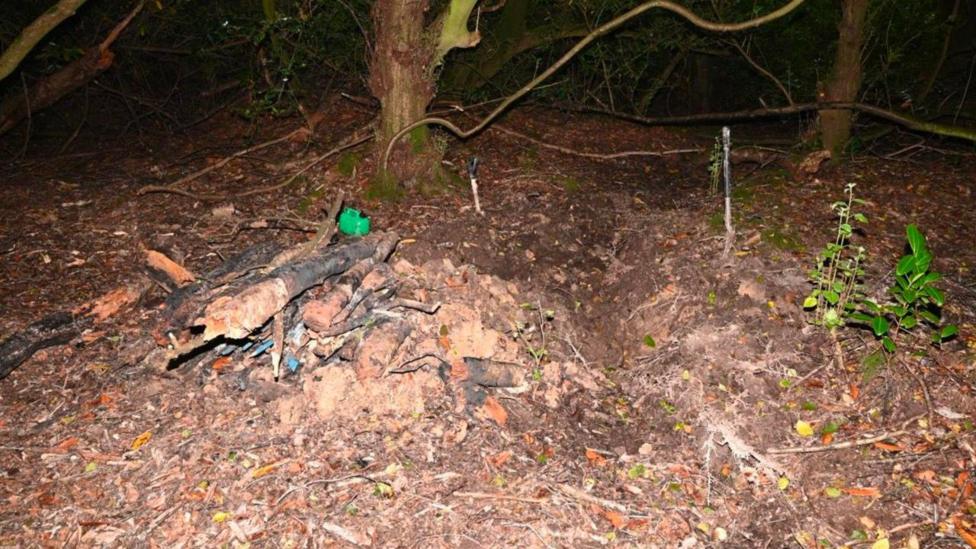 Image taken at night with some kind of artificial lighting or flash lighting it. Shows two tree trunks in the background and a forest floor. Towards the middle of the image is a pile of logs and other material. A green petrol can is also visible and what appears to be an indentation in the ground to the right of the pile of logs.