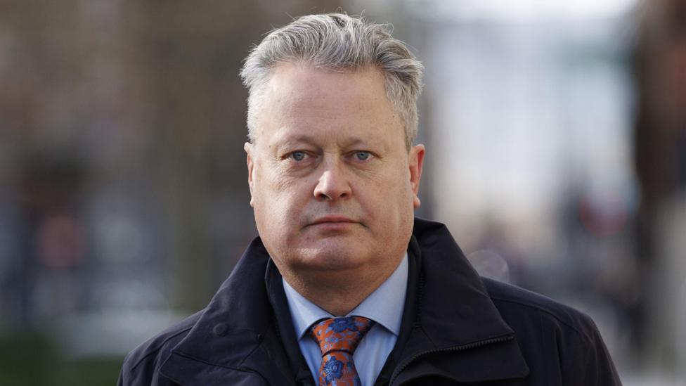 Vincent Kearney is looking directly at the camera on an outside street. The background to the image is out of focus. Vincent has short grey hair and is wearing a navy trenchcoat, blue shirt and colourful red tie, which is covered in blue flowers.