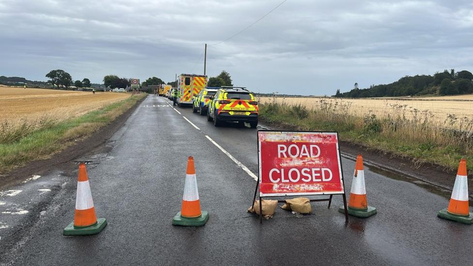 A road closed sign on a country road with a large number of emergency services vehicles in the background