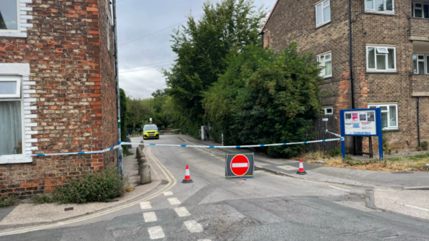 A street with what appears to be residential buildings on either side. There is blue and white police tape cordoning off access to the area and a red circle stop sign placed in the road.