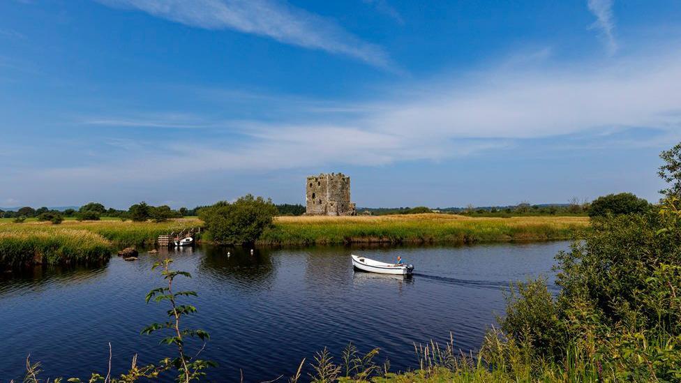 A small boat in the River Dee halfway across to the island on which the imposing Threave Castle stands