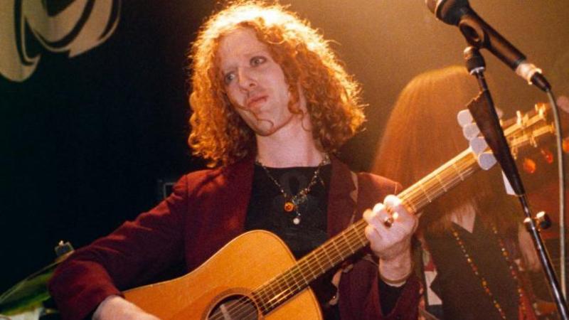 Glen Hansard has long red, curly hair and seems to be wearing eye makeup and is playing a guitar