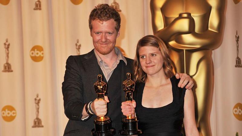 Glen Hansard and Markéta Irglova hold their Oscars. Hansard has short red hair and stubble and is wearing a suit jacket and open-necked shirt. Irglova is wearing a black dress and has shoulder-length reddish/brown hair