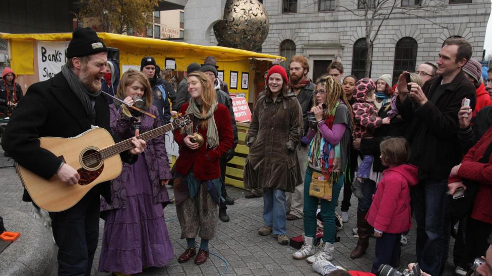 Glen Hansard wearing a beanie hat and coat busks in Dublin watched by a number of people
