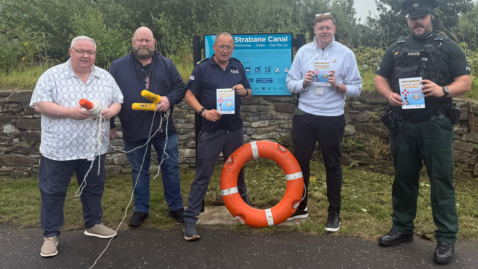 Five men, including on the far right a police officer in unifrom, stand in front of a sign saying Strabane Canal - behind the sign sits the canal itself. Each of the men are holding life saving equipoment