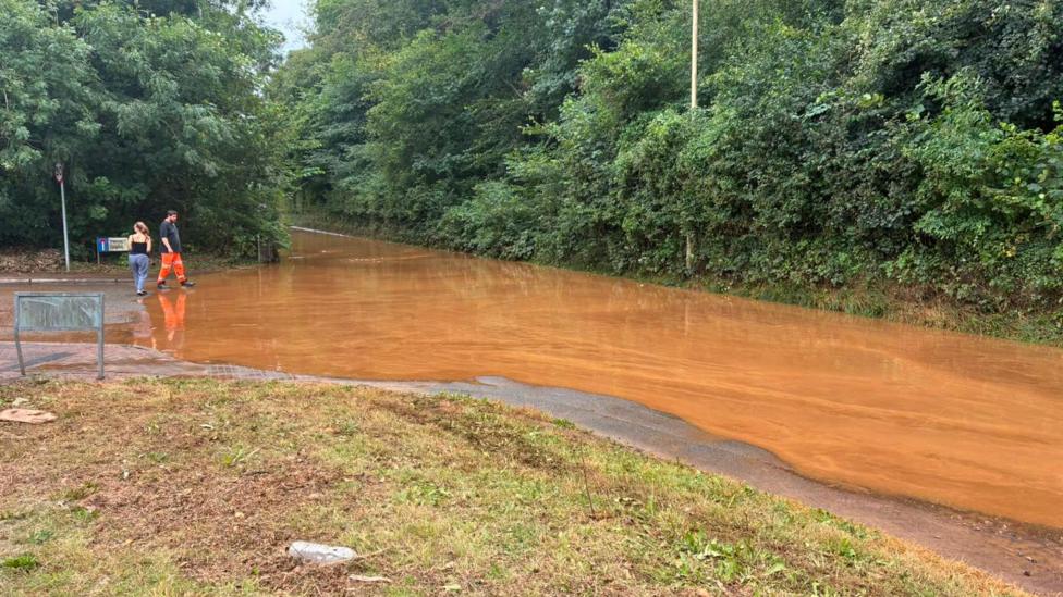 Flooding at Gwaelod-y-Garth, Taff's Well, Cardiff shows a road full of mud and stones, with brown water covering the road.