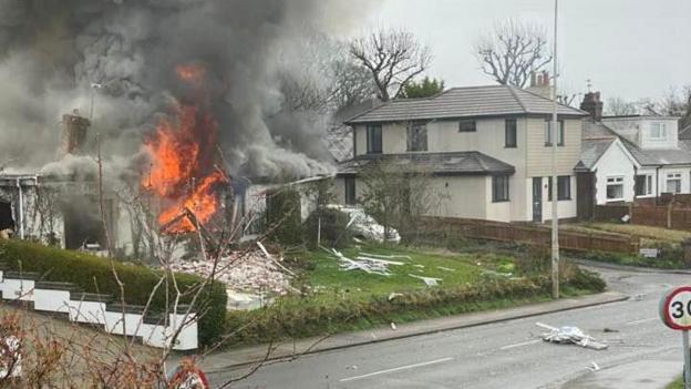 House in flames on a street - billowing grey smoke rises from the single-storey white building