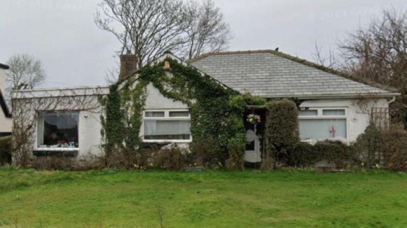 A white-rendered bungalow, with a grey tiled roof and ivy growing around one window and the door, sits on a plot of green grass