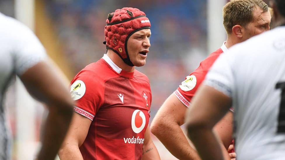 James Botham in a Wales shirt. He wears a red protective cap, and stands with other players on the pitch.
