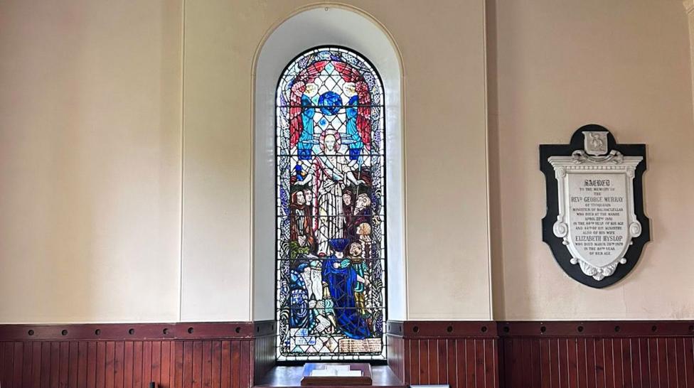 A more distant view of a stained-glass window in a rural south of Scotland church featuring Jesus at its centre and people gathered in reverence around