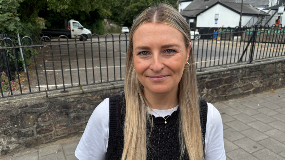 Beth is pictured wearing a black top and a white t-shirt underneath. She is looking at the camera and smiling, and behind her is a car park.