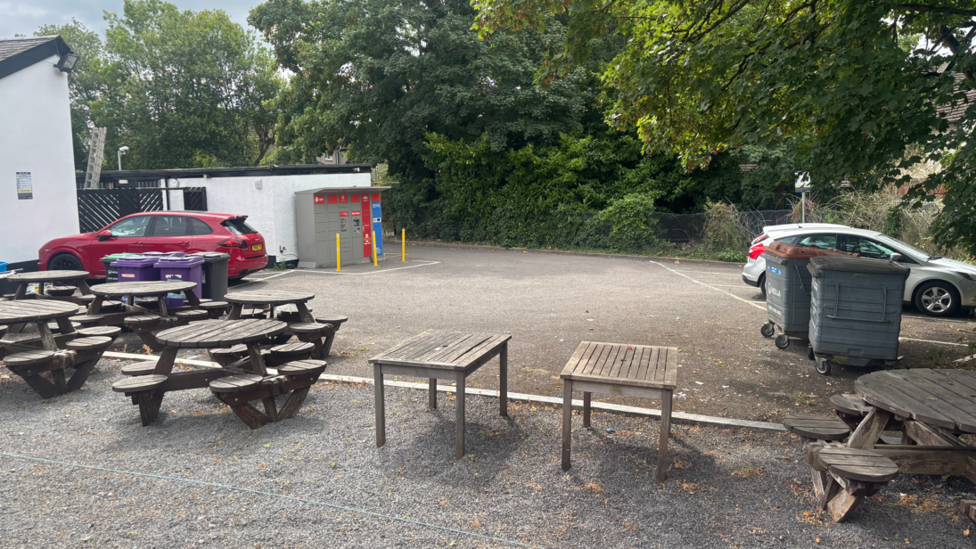 A small car park is pictured with some cars parked in spaces, as well as benches. In the background is a parcel locker.