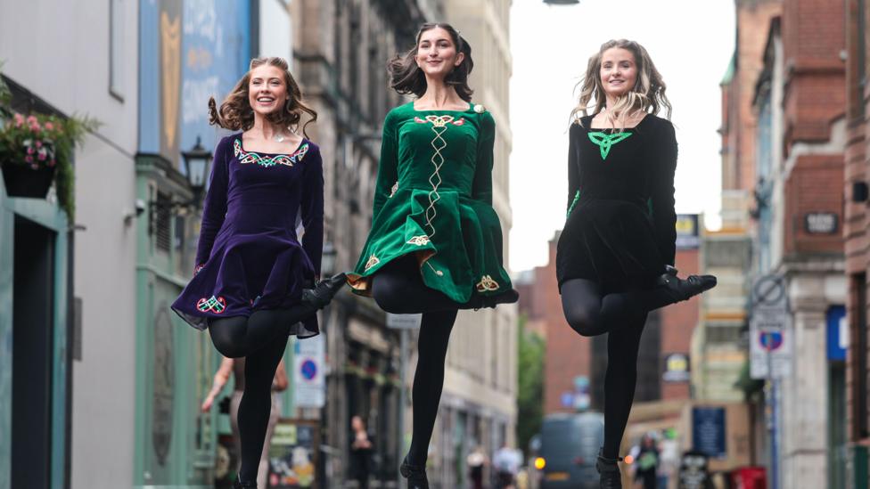 Three women in traditional Irish dancing dresses - jewel toned velvet with celtic embellishments. They are pictured dancing, mid leap in the air on a city street. 