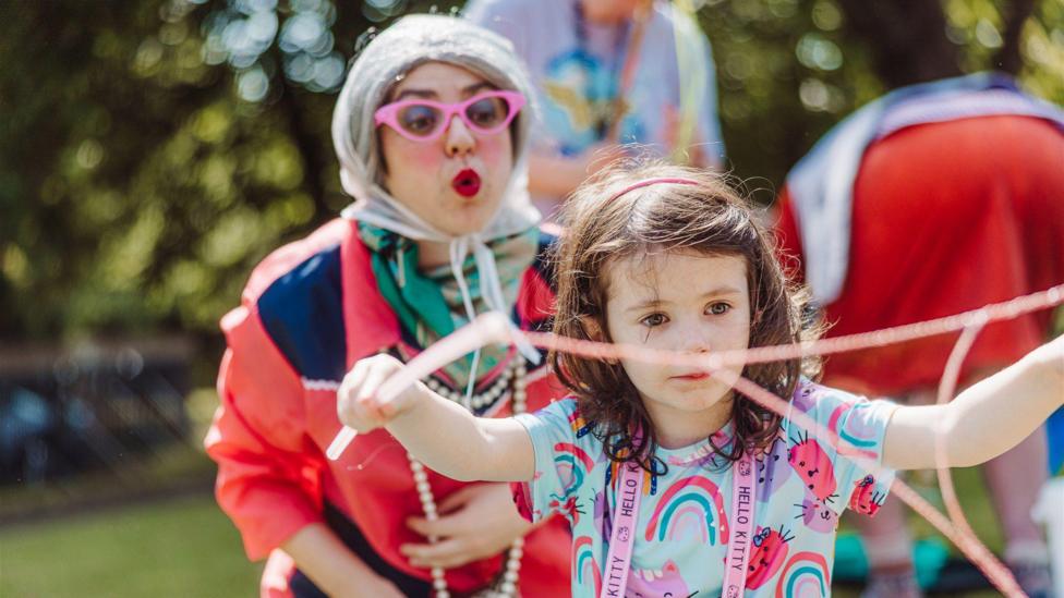 A woman wearing pink glasses, a colourful green scarf, red jacket and bonnet, blowing bubbles toward a young brown haired girl who is playing with a streamer or similar. They are outside at a street party environment. 