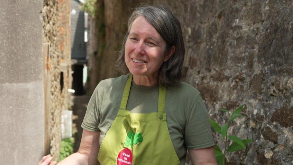 Chantelle Norton, a director of Food in Community CIC, stands smiling in front of a wall covered with lichen and moss. She is wearing an olive green apron bearing the words