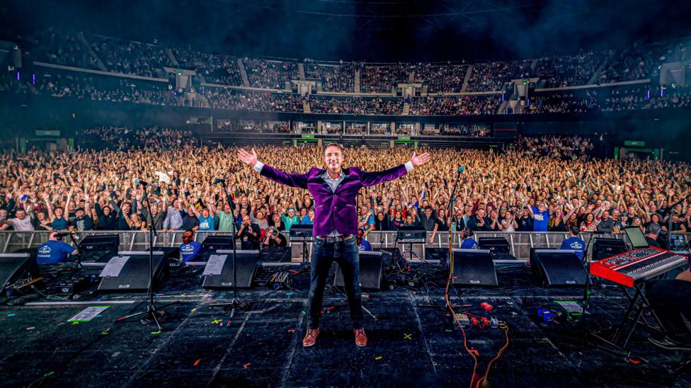 Gary Innes, a man wearing a purple jacket and jeans, standing on stage at the OVO Hydro during a previous Hoolie in the Hydro event. A crowd of thousands, cheering and waving, are behind him.
