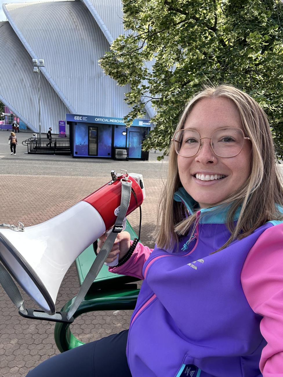 Alice McNaugher, sitting in a tall chair similar to those used by tennis umpires. She is wearing a purple and pink jacket and holding a megaphone in one hand. She is sitting outside at the SEC campus in Glasgow.