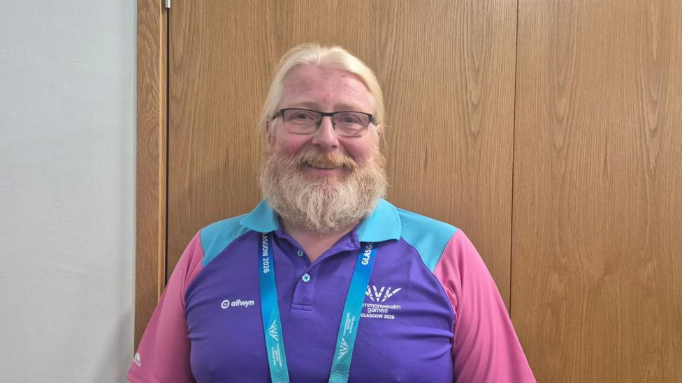 Peter Craig - a man with a fair beard, glasses and fair hair, wearing a commonwealth games uniform of purple, blue and pink.