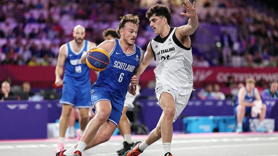 Kyle Jimenez of Team Scotland - wearing an all blue basketball uniform - is challenged by Christian Martin of Team New Zealand, wearing all white. Other players can be seen in the background, while the crowd are blurry in the distance.