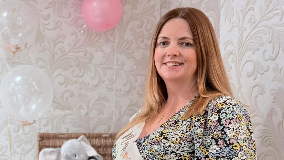 A pregnant woman at a baby show with a floral dress on. A framed picture and a hamper with toys is in the background