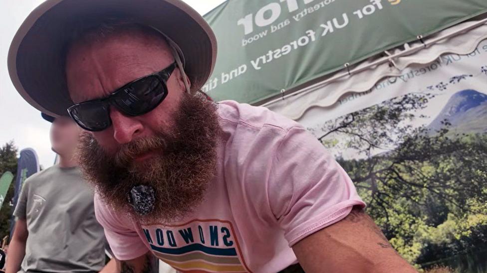 A man with a bushy beard, wearing a pink T shirt, looks at the camera