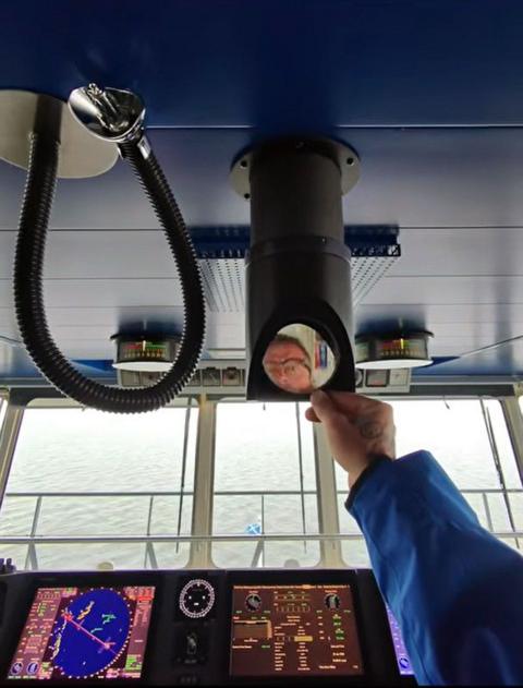 A man wearing glasses looks into a small mirror on a piece of equipment in the bridge of a large ferry