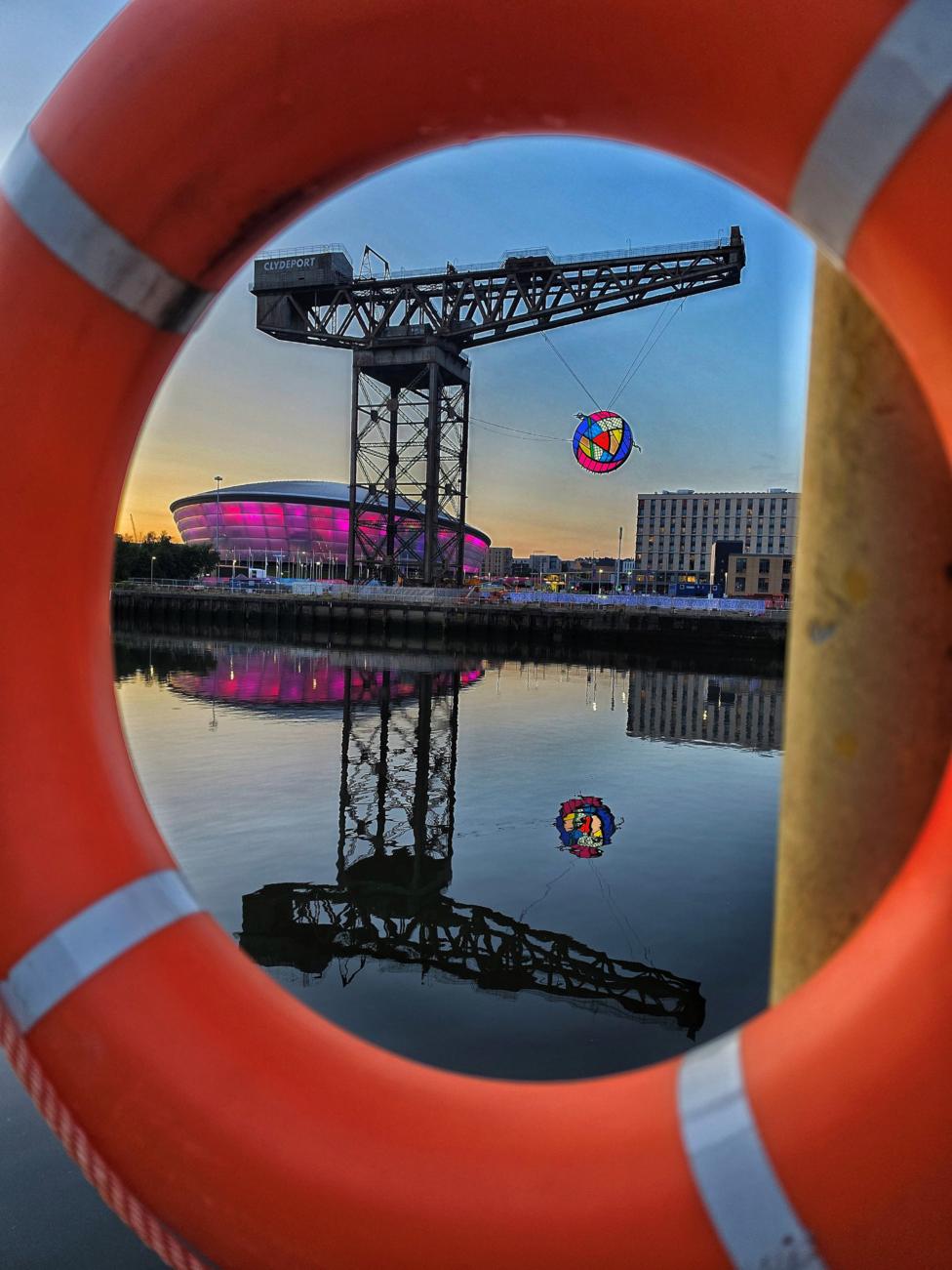 A large orange lifebuoy frames a view across the River Clyde. Through the centre of the ring, the Finnieston crane can be seen with the Commonwealth Games rose sculpture raised in the air and the Hydro illuminated in pink in the background.