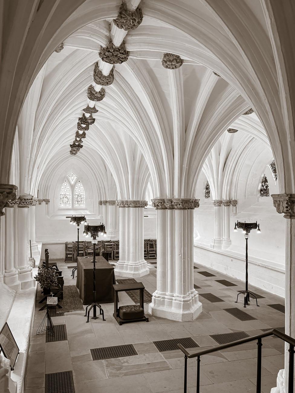 An ornate stone hall with soaring Gothic vaulted ceilings and carved columns stretches into the distance. Decorative stone bosses line the ceiling, while tables, chairs and tall lamps sit beneath arched windows that let light into the historic interior.