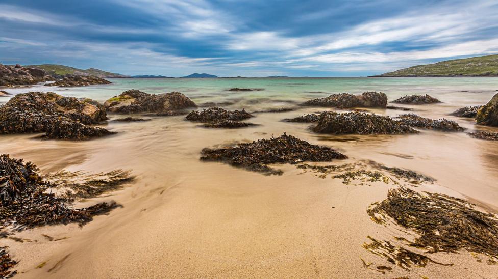 Seaweed-covered rocks dot a shallow bay with pale sand and turquoise-green water. The long-exposure photograph creates a smooth, flowing effect in the water beneath dramatic blue-grey clouds and distant low hills.